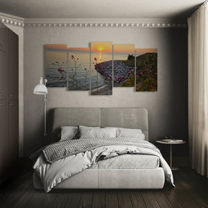 A multi-panel canvas wall art set depicting a coastal sunset landscape with pink flowers, displayed above a gray upholstered bed in a modern bedroom setting.