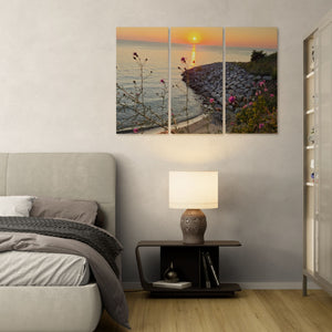 A three-panel canvas wall art print depicting a sunset over a rocky shoreline with water and flowers, displayed in a modern bedroom setting above a bed and next to a nightstand.