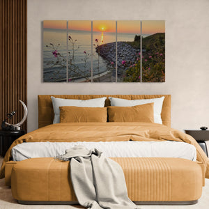 A five-panel canvas wall art print depicting a coastal sunset over the ocean, styled in a modern bedroom above a bed with mustard yellow bedding and a matching bench.