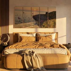 A staged bedroom scene with a large multi-panel wall art print showing a beach shoreline at sunset, hung above a bed with tan bedding and matching furniture in warm golden light.