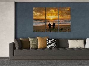 Three-panel artwork of a sunset over the ocean with people on a beach, displayed above a gray sofa with patterned cushions.